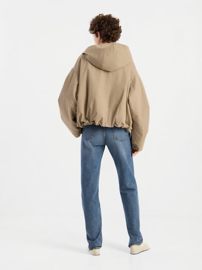 Eine Person mit lockigem Haar trägt die Jacke „Savanna“, eine übergroße beige Damenjacke mit Kapuze, dazu blaue Jeans und weiße Schuhe. Sie steht vor einem schlichten weißen Hintergrund und ist mit dem Rücken zur Kamera gewandt.