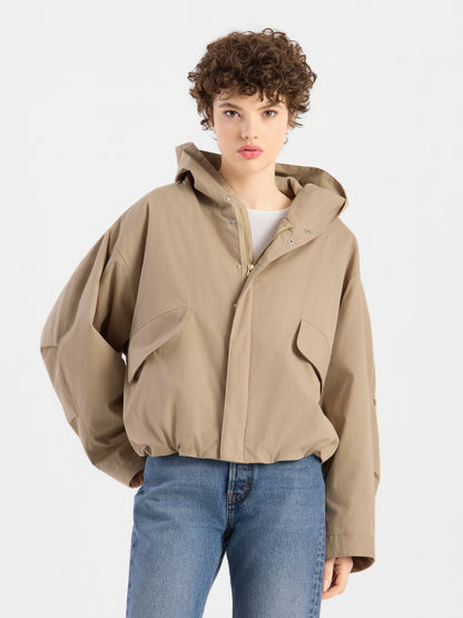 Eine Person mit kurzen Locken trägt die hellbraune Jacke „Savanna“ mit Kapuze und eine blaue Jeans; sie steht vor einem schlichten weißen Hintergrund, hat einen neutralen Gesichtsausdruck und steckt eine Hand in die Hosentasche.