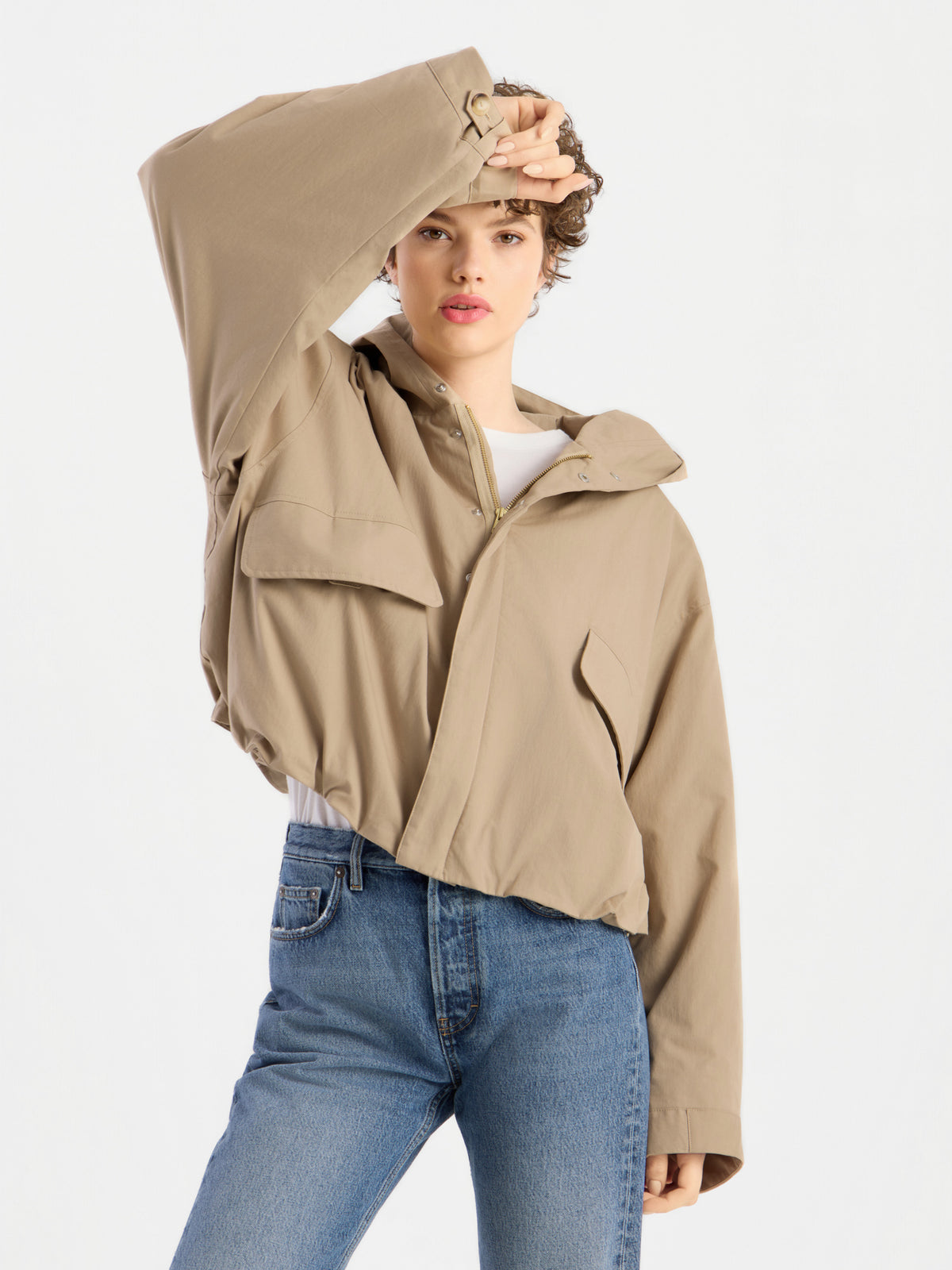 Eine Person, die die Jacke „Savanna“ in Hellbraun mit Kapuze trägt und eine blaue Jeans, posiert mit einem erhobenen Arm, wobei die Hand ihr Gesicht teilweise verdeckt, vor einem schlichten weißen Hintergrund.