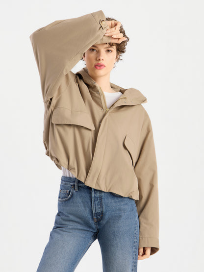 Eine Person, die die Jacke „Savanna“ in Hellbraun mit Kapuze trägt und eine blaue Jeans, posiert mit einem erhobenen Arm, wobei die Hand ihr Gesicht teilweise verdeckt, vor einem schlichten weißen Hintergrund.