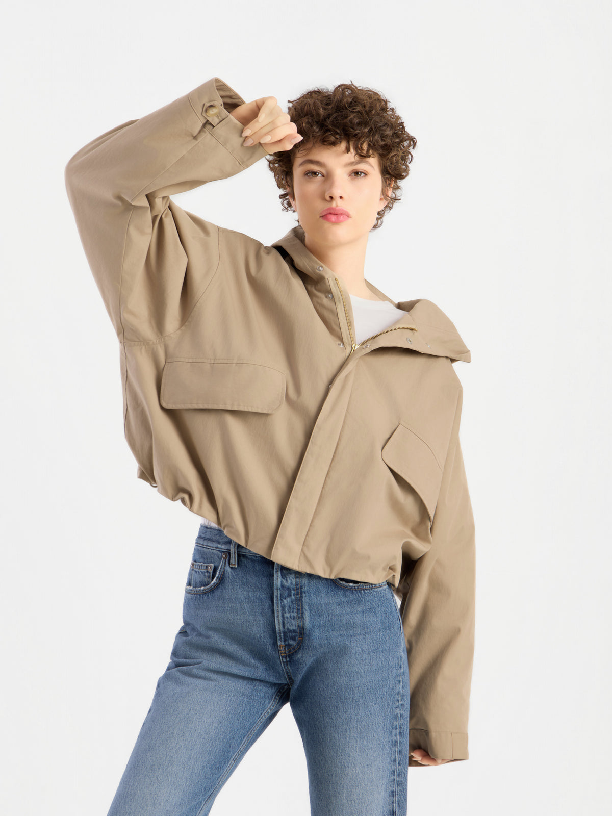 Eine Person mit kurzen Locken posiert selbstbewusst in der beigen Jacke „Savanna“ und blauen Jeans, hebt einen Arm und blickt vor einem schlichten weißen Hintergrund in die Kamera.
