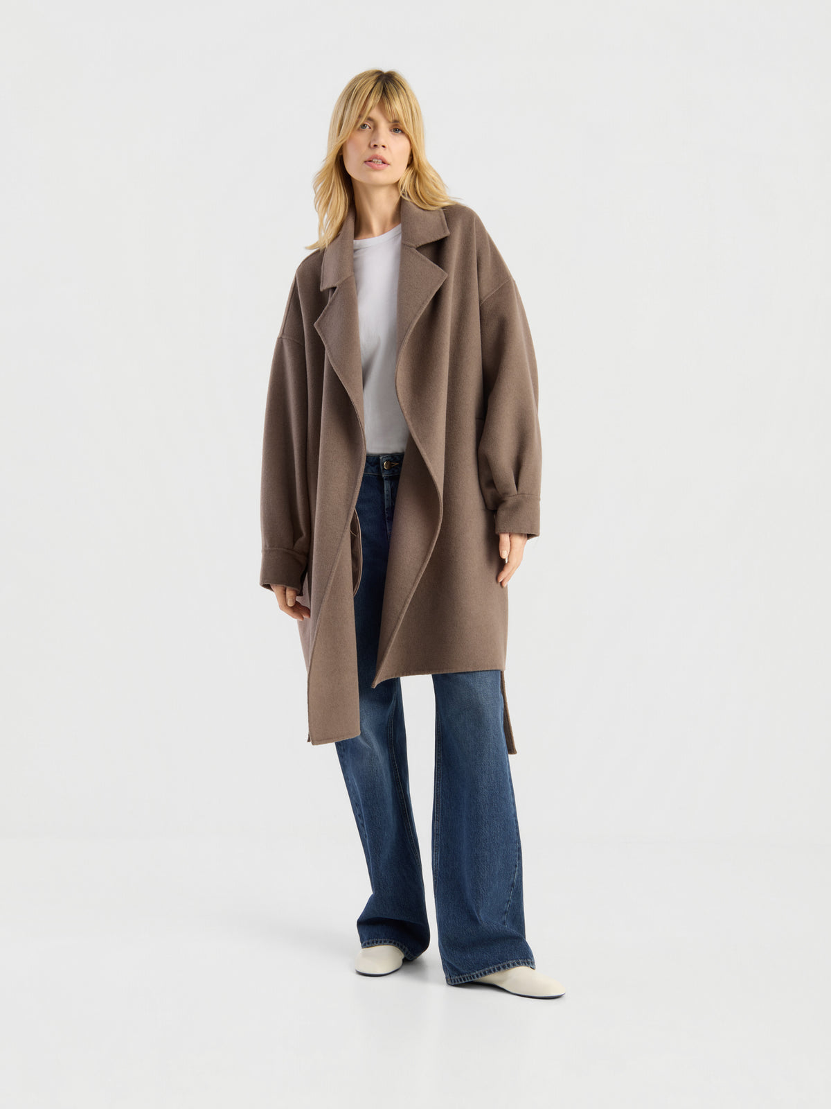 Eine Person, die den „Mantel Francesca Taupe“ trägt – einen Oversize-Mantel in einem hellen Braunton –, der über einem weißen Hemd, einer blauen Hose mit weitem Bein und weißen Schuhen getragen wird, steht vor einem schlichten weißen Hintergrund.