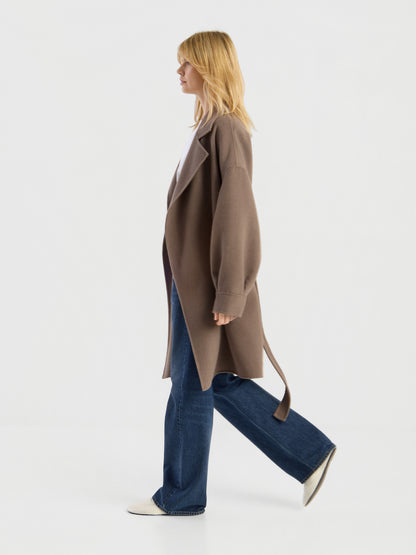 Eine Frau in einem Mantel von Francesca Taupe, einem weißen Hemd und blauen Jeans, die vor einem schlichten weißen Hintergrund nach links geht.