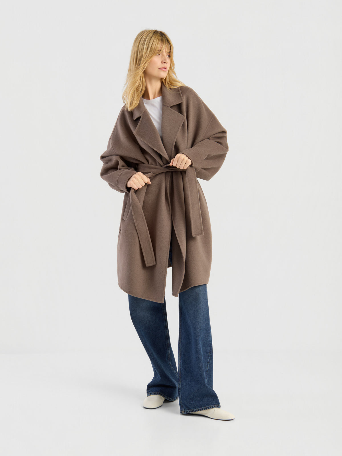 Vor einem schlichten weißen Hintergrund steht eine Frau mit blonden Haaren. Sie trägt den eleganten Mantel „Francesca Taupe“ über einem weißen Oberteil, einer blauen Jeans mit weitem Bein und weißen Schuhen.