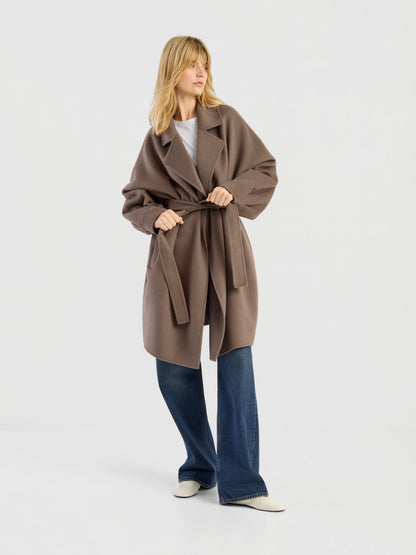 Vor einem schlichten weißen Hintergrund steht eine Frau mit blonden Haaren. Sie trägt den eleganten Mantel „Francesca Taupe“ über einem weißen Oberteil, einer blauen Jeans mit weitem Bein und weißen Schuhen.