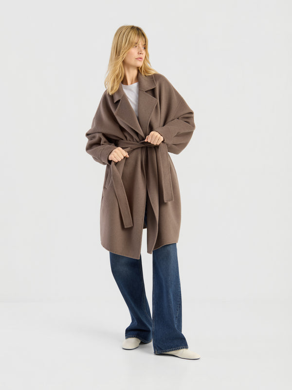 Eine Frau, die den langen Mantel „Cozy“ in Taupe mit Gürtel über einem weißen Oberteil, einer blauen Jeans mit weitem Bein und weißen Schuhen trägt, steht vor einem schlichten weißen Hintergrund.