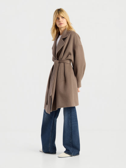 Eine Person, die den eleganten Mantel „Francesca Taupe“ trägt, der an der Taille mit einem Gürtel über einer blauem Jeans mit weitem Bein und weißen Schuhen gebunden ist, steht vor einem schlichten weißen Hintergrund und blickt leicht zur Seite.
