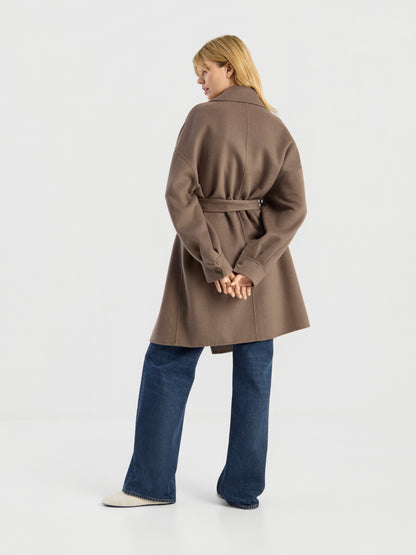 Eine Frau steht mit dem Rücken zur Kamera vor einem schlichten weißen Hintergrund. Sie trägt den Mantel „Francesca Taupe“ von Mantel mit Gürtel, eine blaue Jeans und weiße Schuhe, wobei sie die Hände hinter dem Rücken verschränkt hat.