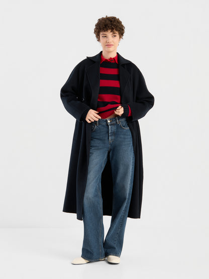 Eine Person mit kurzen Locken trägt den Mantel „Audrey Dunkelblau“, einen rot-schwarz gestreiften Pullover, blaue Jeans mit weitem Bein und weiße Schuhe und steht vor einem schlichten weißen Hintergrund.