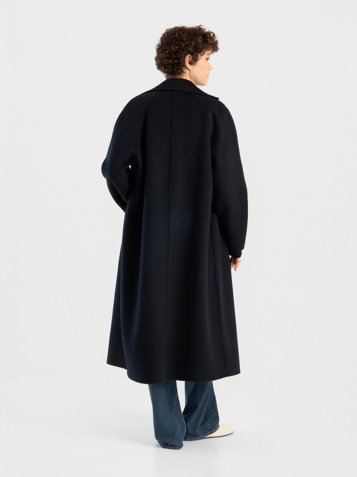 Eine Person mit kurzen Locken, die den Mantel „Audrey Dunkelblau“, eine blaue Jeans und weiße Schuhe trägt, steht mit dem Rücken zur Kamera in einem hellen, minimalistisch gestalteten Studio.