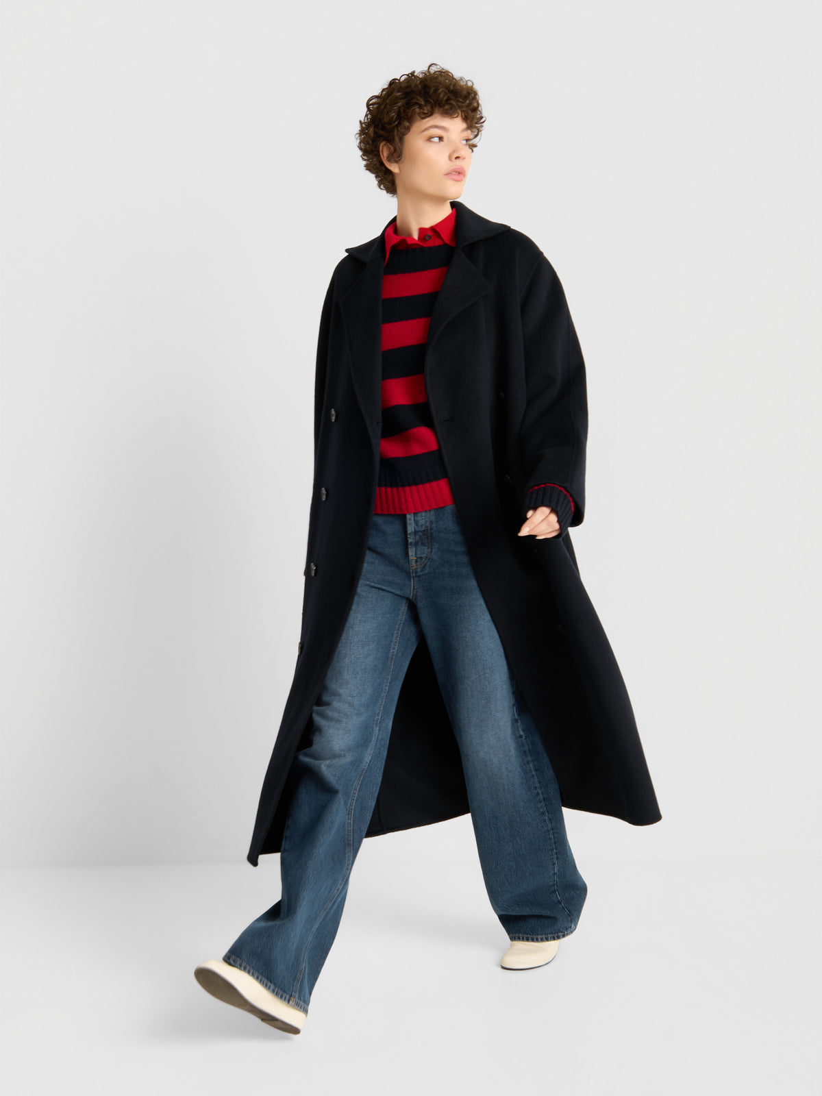 Eine Person, die den „Mantel Audrey Dunkelblau“, einen rot-schwarz gestreiften Pullover, eine Jeans mit weitem Bein und weiße Schuhe trägt, steht vor einem schlichten, hellen Hintergrund und blickt nach rechts.