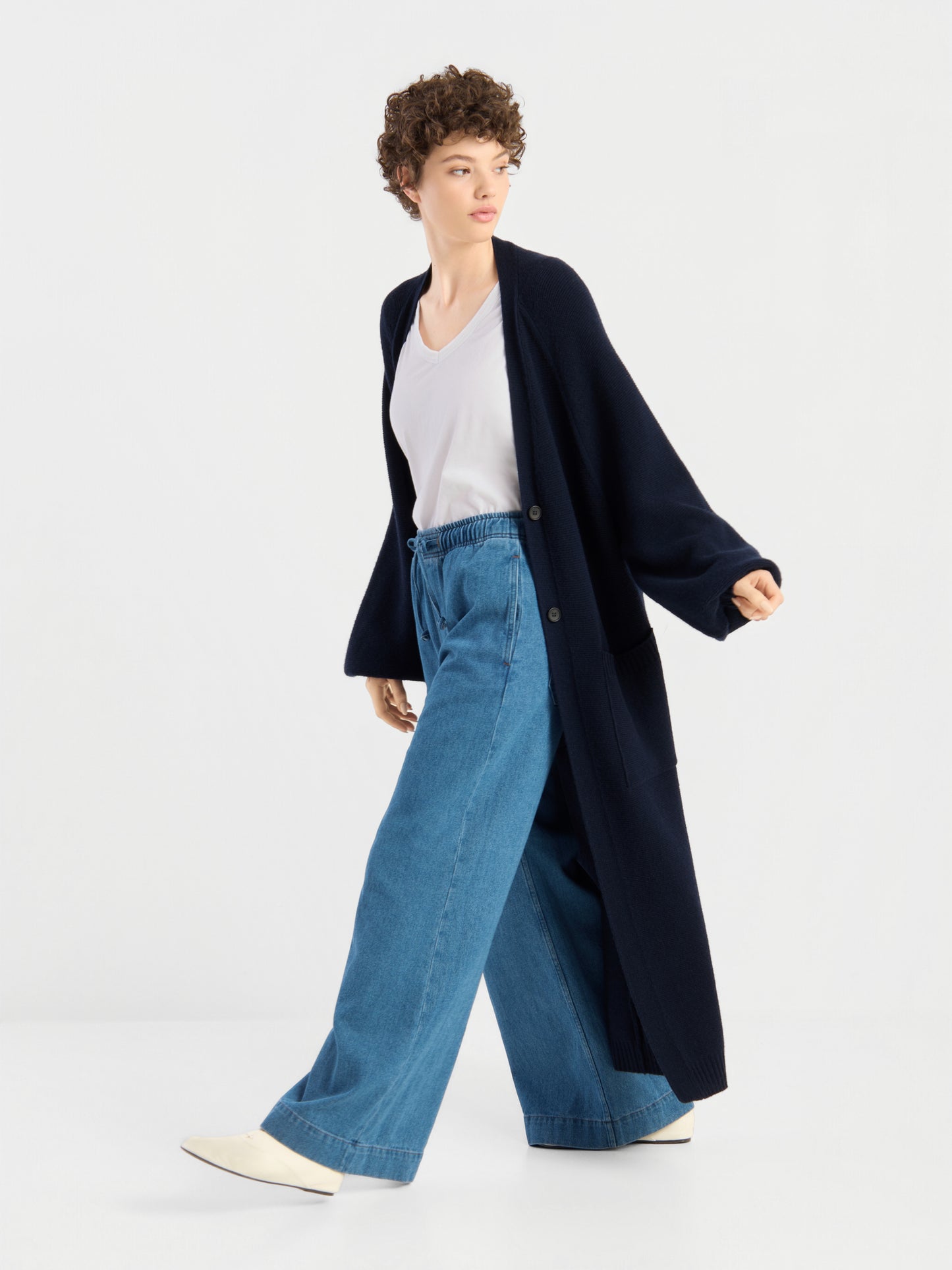 Eine Person mit kurzen Locken, die den „Mantel Antonia Vol. II“ trägt – eine lange, dunkelblaue Strickjacke aus weicher Merinowolle –, kombiniert mit einem weißen T-Shirt, einer blau-weit geschnittenen Jeans und weißen Schuhen, geht vor einem schlichten weißen Hintergrund.