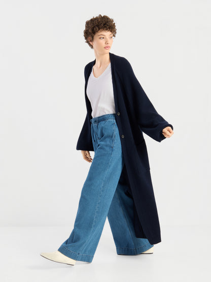 Eine Person mit kurzen Locken, die den „Mantel Antonia Vol. II“ trägt – eine lange, dunkelblaue Strickjacke aus weicher Merinowolle –, kombiniert mit einem weißen T-Shirt, einer blau-weit geschnittenen Jeans und weißen Schuhen, geht vor einem schlichten weißen Hintergrund.