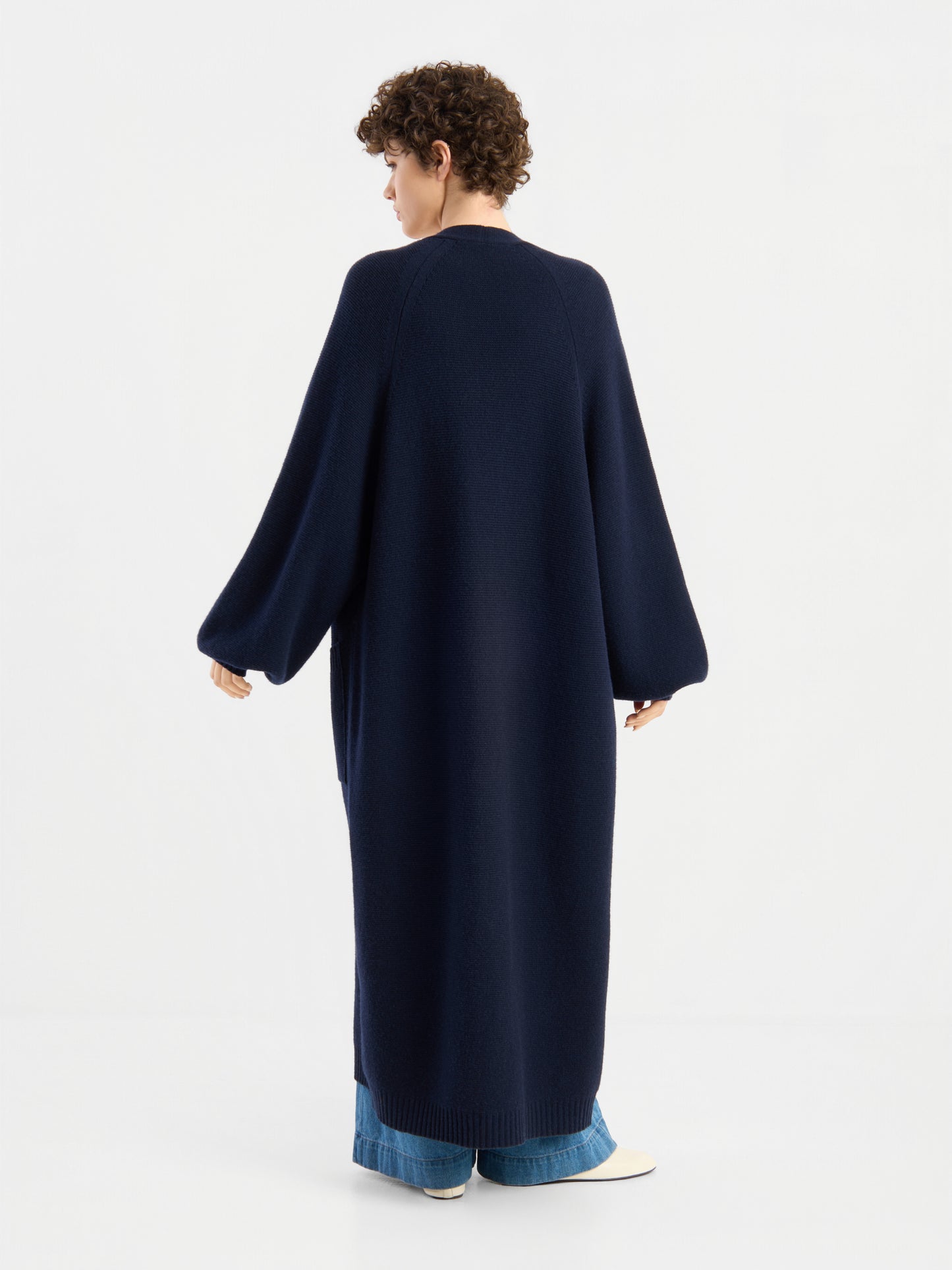 Eine Person mit kurzen Locken trägt den langen, marineblauen Mantel „Antonia Vol. II“ über einer blauen Jeans und weißen Schuhen; sie steht vor einem schlichten weißen Hintergrund und ist mit dem Rücken zur Kamera gewandt.