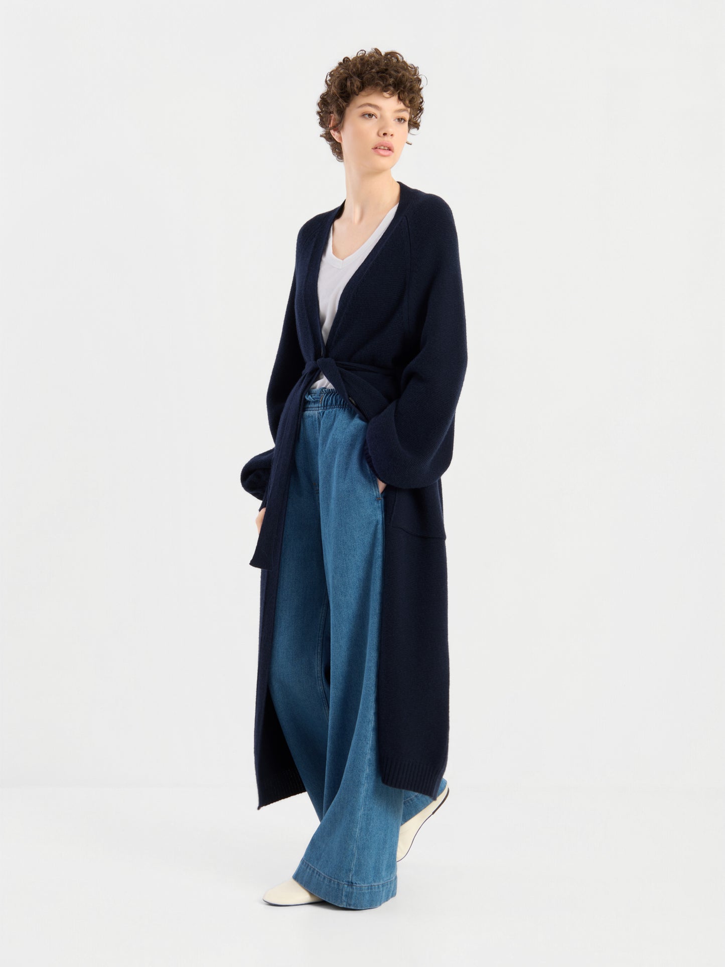 Eine Person mit kurzen Locken trägt den langen, marineblauen Mantel „Antonia Vol. II“ aus Merinowolle über einem weißen Oberteil und einer blau-weit geschnittenen Jeans, dazu weiße Schuhe, und steht vor einem schlichten weißen Hintergrund.