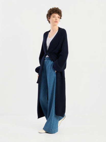 Eine Person mit kurzen Locken trägt den langen, marineblauen Mantel „Antonia Vol. II“ aus Merinowolle über einem weißen Oberteil und einer blau-weit geschnittenen Jeans, dazu weiße Schuhe, und steht vor einem schlichten weißen Hintergrund.