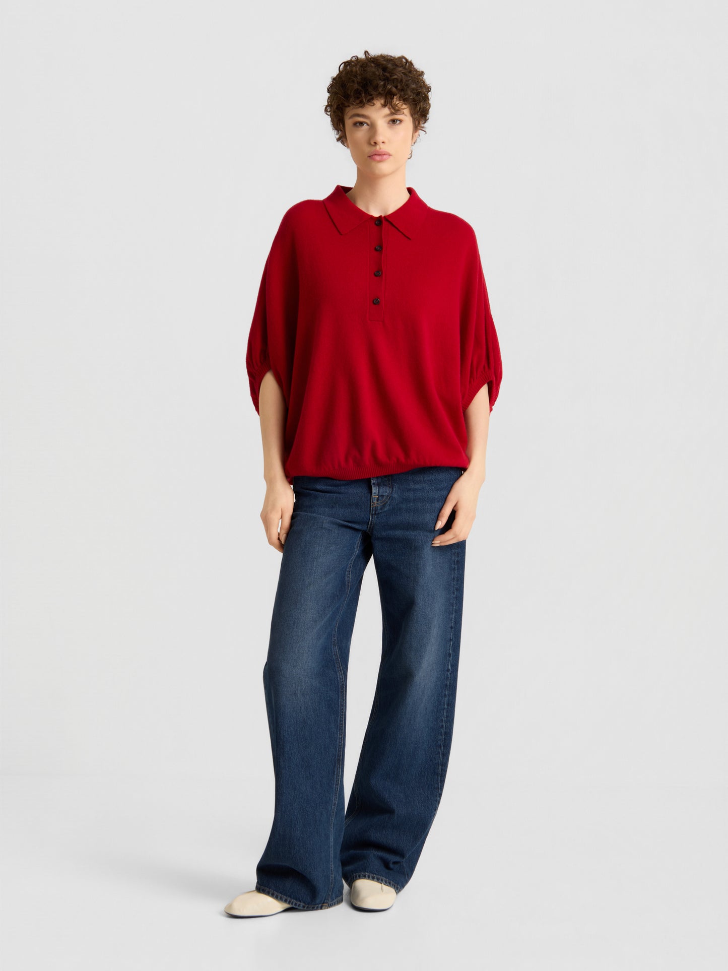 Eine Person mit kurzen, lockigen Haaren und einem ausdruckslosen Gesichtsausdruck steht vor einem schlichten Hintergrund und trägt das „Cowboy Polo Knit Red“, eine blaue Jeans mit weitem Bein und weiße Schuhe.