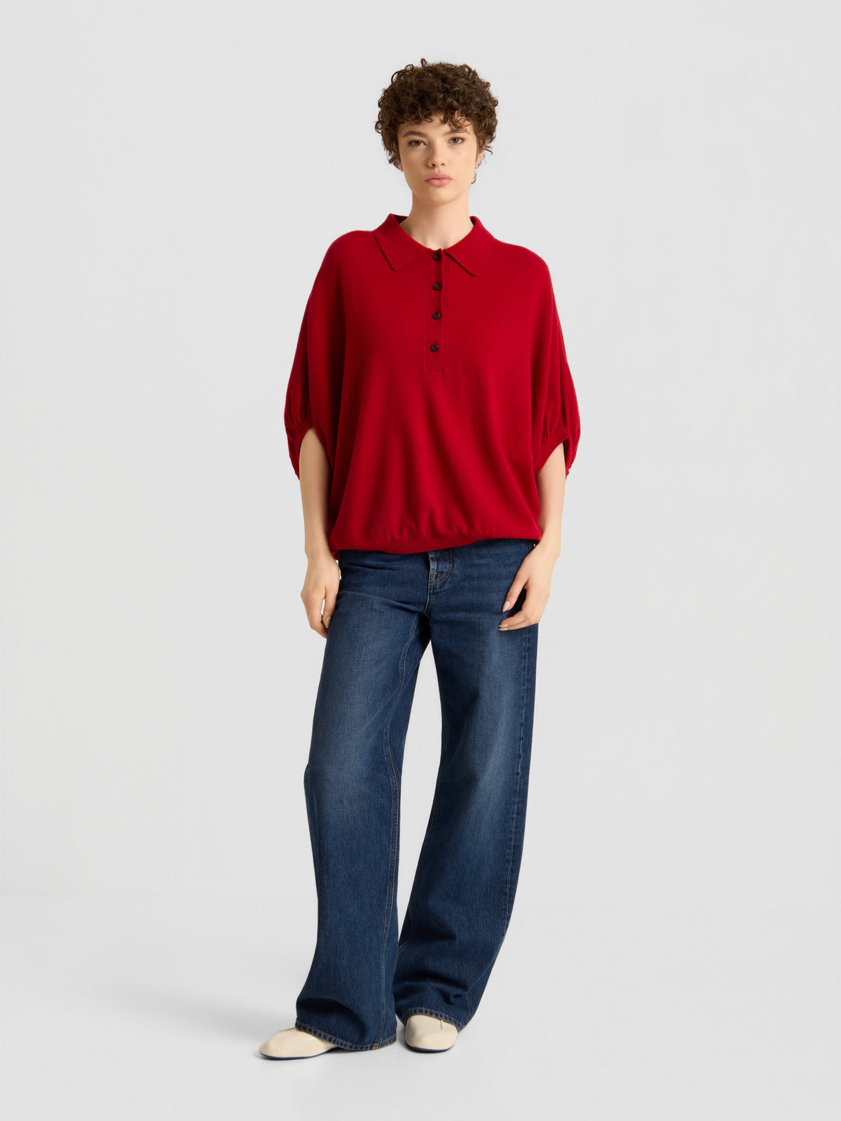 Eine Person mit kurzen, lockigen Haaren und einem ausdruckslosen Gesichtsausdruck steht vor einem schlichten Hintergrund und trägt das „Cowboy Polo Knit Red“, eine blaue Jeans mit weitem Bein und weiße Schuhe.