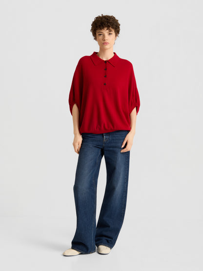 Eine Person mit kurzen, lockigen Haaren und einem ausdruckslosen Gesichtsausdruck steht vor einem schlichten Hintergrund und trägt das „Cowboy Polo Knit Red“, eine blaue Jeans mit weitem Bein und weiße Schuhe.