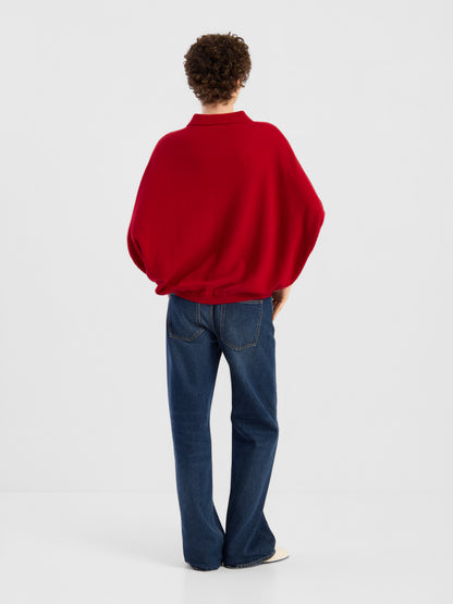 Eine Person mit kurzen Locken, die ein rotes „Cowboy Polo Knit“-Shirt und eine blaue Jeans trägt, steht mit dem Rücken zur Kamera vor einem schlichten weißen Hintergrund.