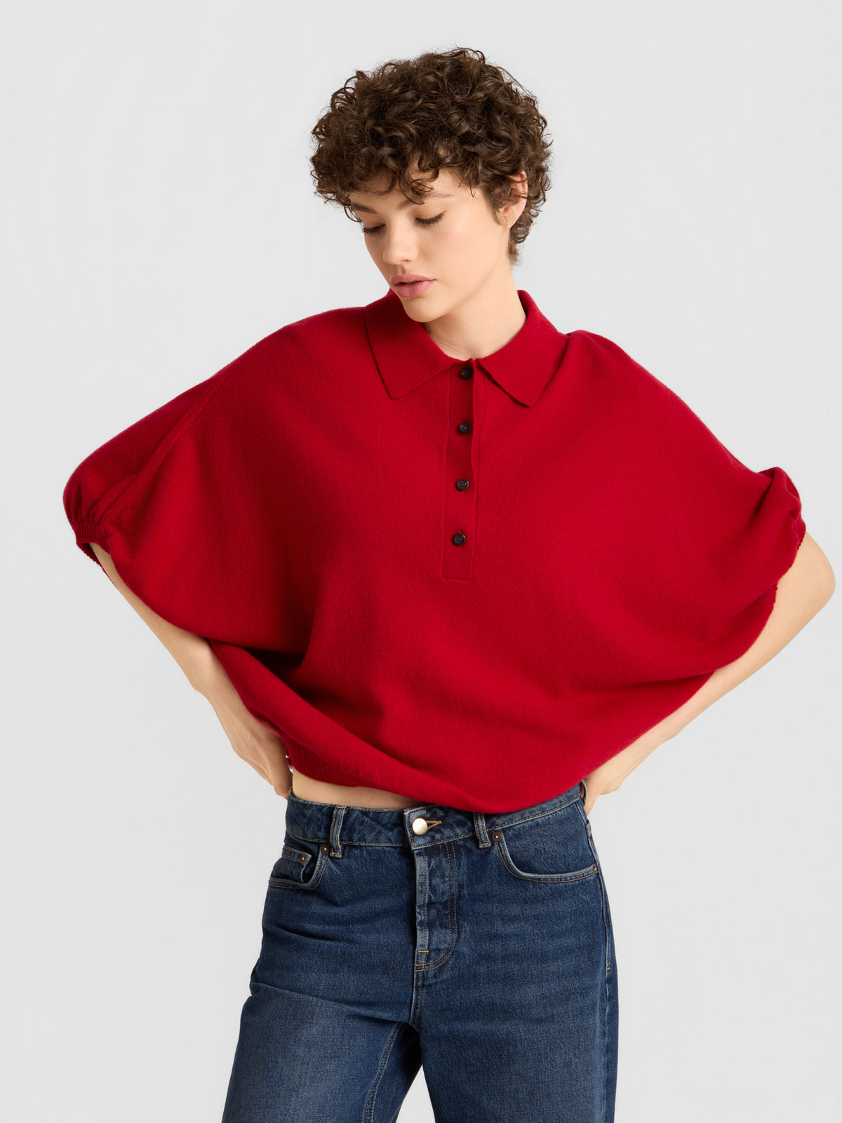 Eine Person mit kurzen Locken trägt das „Cowboy Polo Knit Red“, ein locker sitzendes Oberteil mit Kragen und schwarzen Knöpfen, dazu eine blaue Jeans. Sie steht vor einem schlichten, hellen Hintergrund und hat die Hände in die Hüften gestemmt.