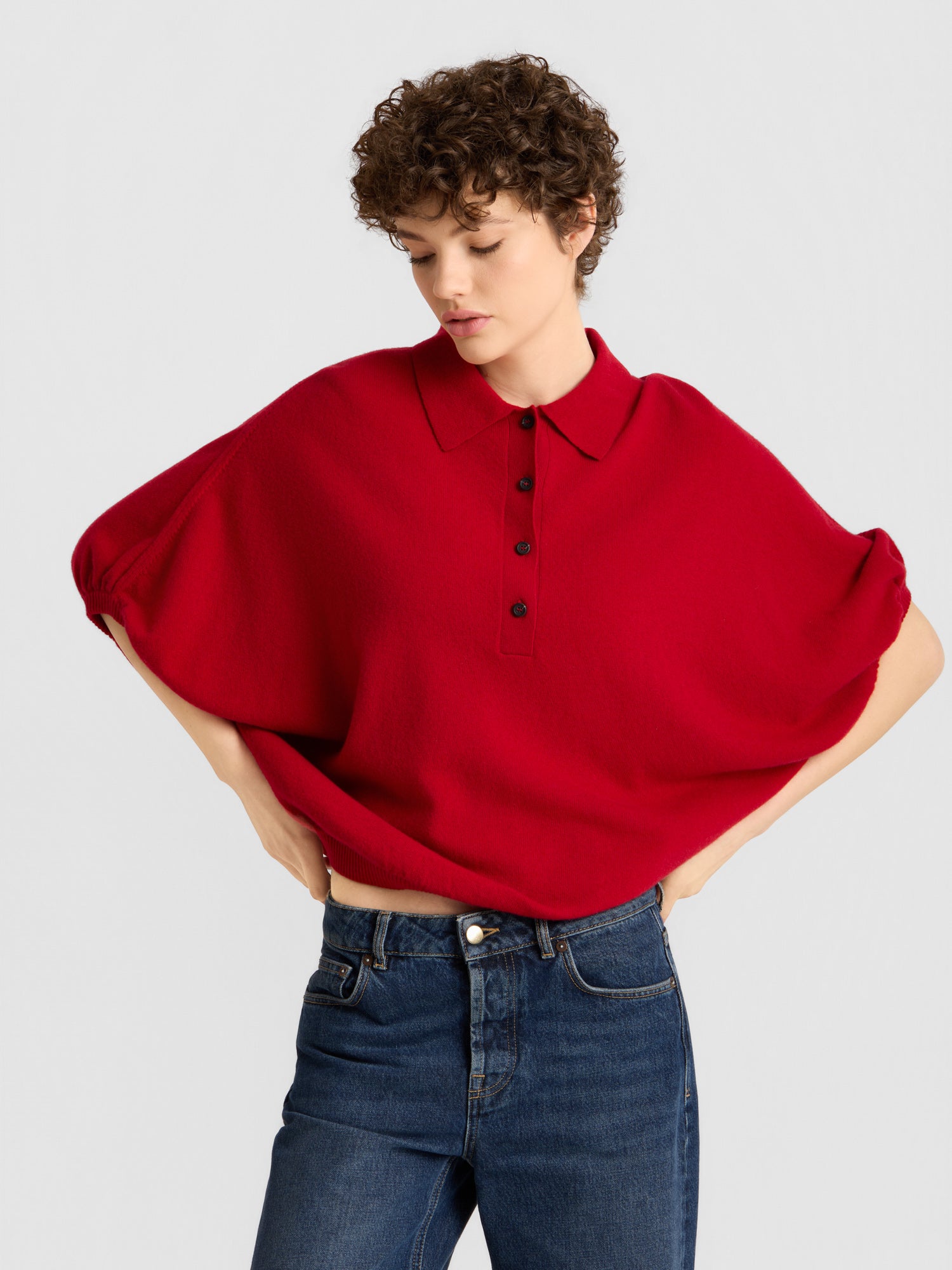 Eine Person mit kurzen Locken trägt das „Cowboy Polo Knit Red“, ein locker sitzendes Oberteil mit Kragen und schwarzen Knöpfen, dazu eine blaue Jeans. Sie steht vor einem schlichten, hellen Hintergrund und hat die Hände in die Hüften gestemmt.