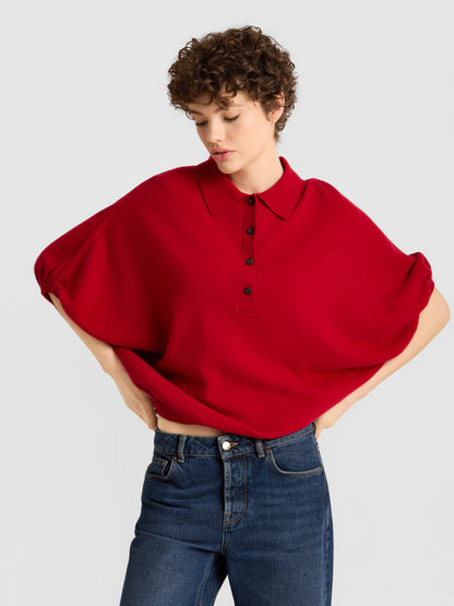 Eine Person mit kurzen Locken trägt das „Cowboy Polo Knit Red“, ein locker sitzendes Oberteil mit Kragen und schwarzen Knöpfen, dazu eine blaue Jeans. Sie steht vor einem schlichten, hellen Hintergrund und hat die Hände in die Hüften gestemmt.