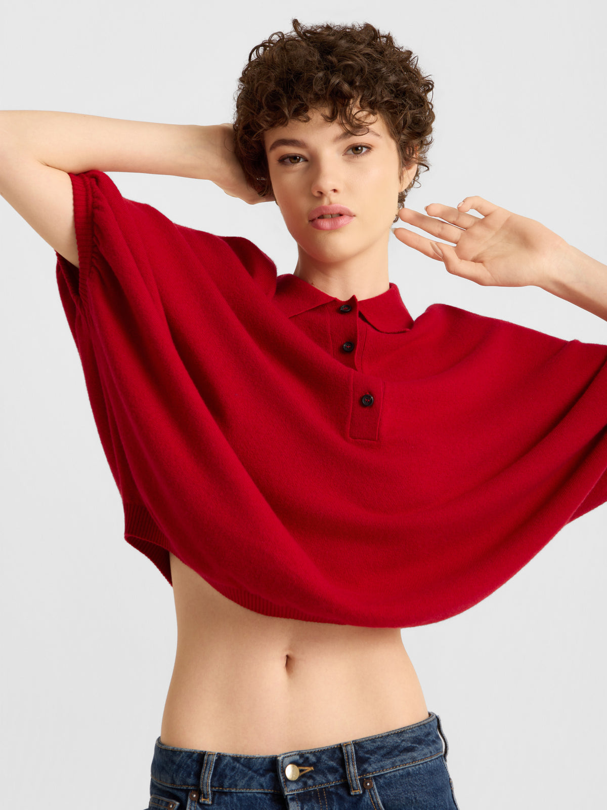 Eine junge Frau mit kurzen Locken präsentiert das „Cowboy Polo Knit Red“, ein leuchtend rotes, locker sitzendes Crop-Top, kombiniert mit einer blauen Jeans. Vor einem schlichten Hintergrund posiert sie selbstbewusst mit einem Arm hinter dem Kopf.