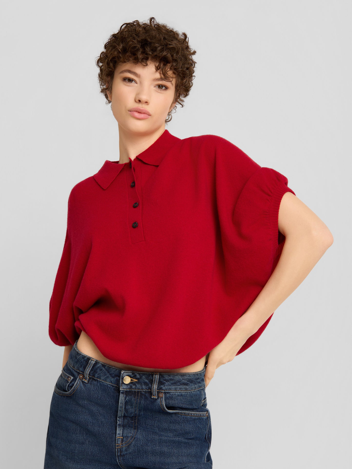 Eine Person mit kurzen Locken trägt das „Cowboy Polo Knit Red“, ein locker sitzendes rotes Oberteil mit Kragen und kurzen Ärmeln, dazu eine dunkelblaue Jeans, und steht vor einem schlichten hellgrauen Hintergrund.