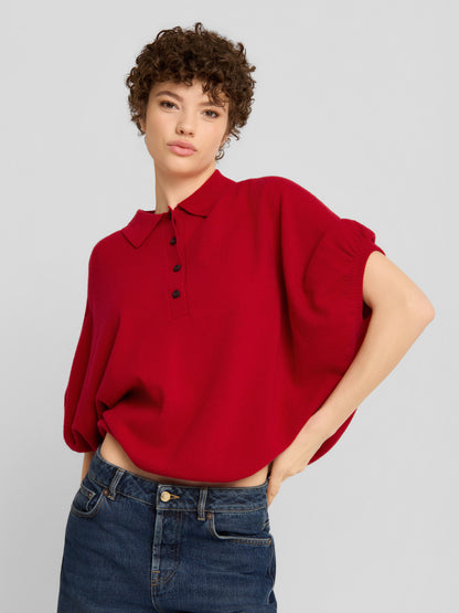 Eine Person mit kurzen Locken trägt das „Cowboy Polo Knit Red“, ein locker sitzendes rotes Oberteil mit Kragen und kurzen Ärmeln, dazu eine dunkelblaue Jeans, und steht vor einem schlichten hellgrauen Hintergrund.