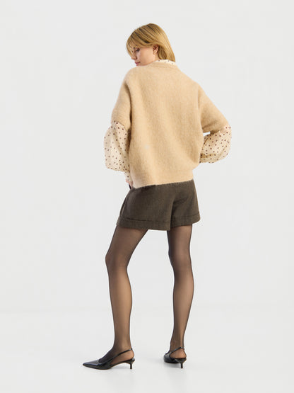 Vor einem schlichten weißen Hintergrund steht eine Frau mit dem Rücken zur Kamera. Sie trägt das „Bear“-T-Shirt in der Farbe „Camel“, dazu dunkle Shorts, schwarze, durchsichtige Strumpfhosen und schwarze Pumps mit Fersenriemen.