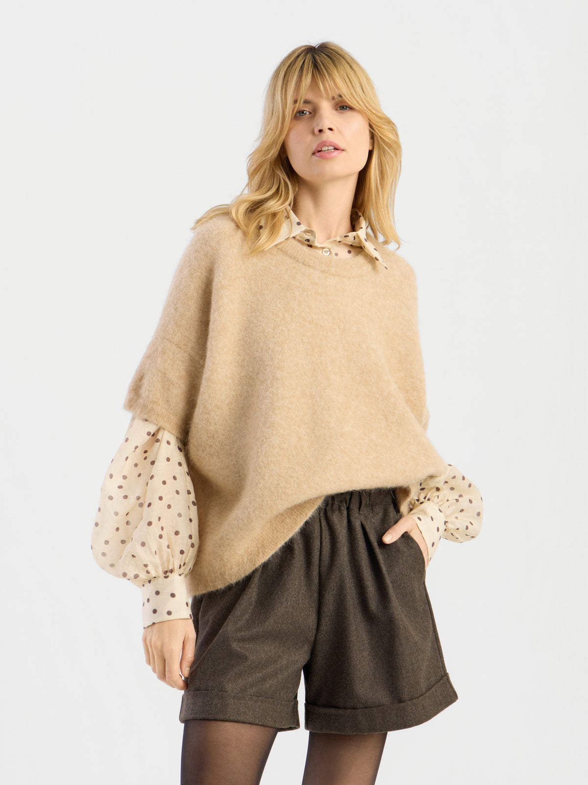 Eine Frau, die das „Bear T-Shirt Camel“ – ein luxuriöses Basic aus Alpaka-Merino – in Beige über einer cremefarbenen Bluse mit Polka-Dots, dunklen Shorts mit hoher Taille und einer schwarzen Strumpfhose trägt, posiert mit einer Hand in der Tasche vor einem schlichten Hintergrund.