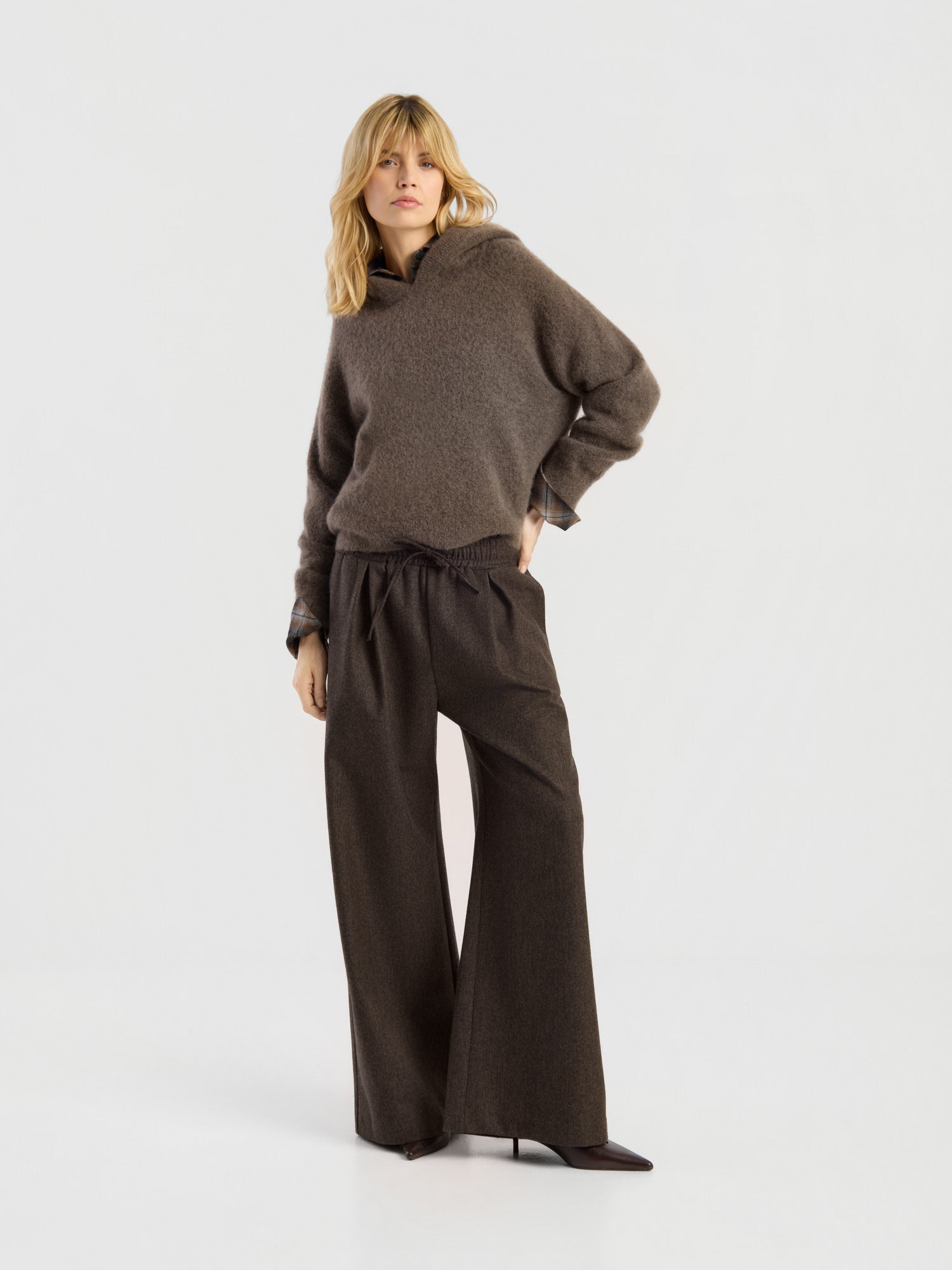 Vor einem schlichten Hintergrund steht eine Frau, die den „Bear Hoodie Braun“ trägt – einen locker sitzenden Damen-Kapuzenpullover aus einer Alpaka-Merino-Wollmischung –, dazu eine braune Hose mit weitem Bein und dunkle, spitze Schuhe. Sie hat eine Hand in die Hüfte gestützt und einen neutralen Gesichtsausdruck.