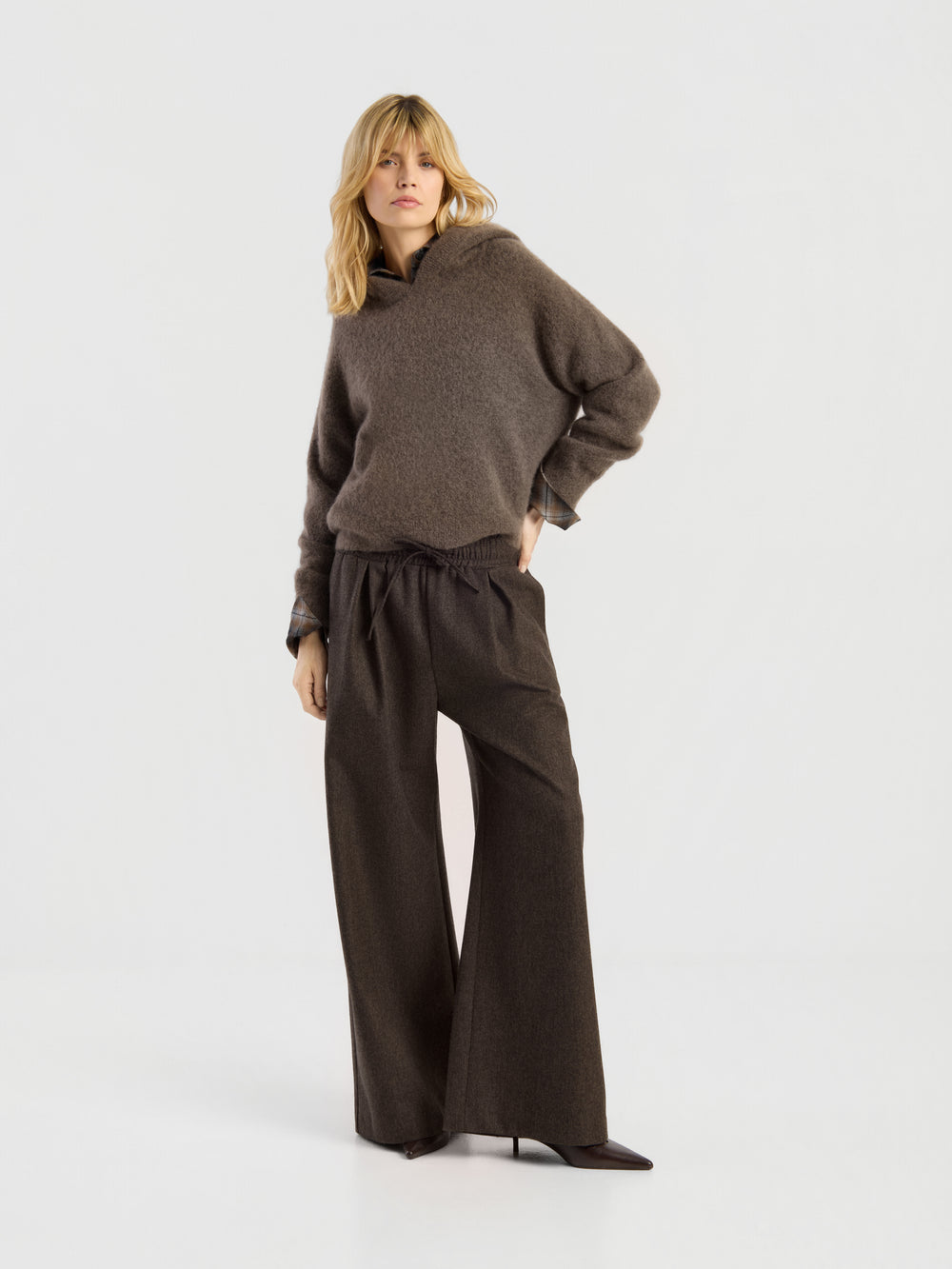 Eine Frau mit blonden Haaren, die einen braunen Pullover mit der Aufschrift „Bear Hug“, eine dunkelbraune Hose mit weitem Bein und Schuhe mit Absatz trägt, steht vor einem schlichten weißen Hintergrund und stützt eine Hand in die Hüfte.