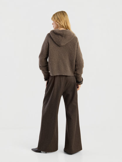 Eine Person mit blonden Haaren, die einen „Bear Hoodie Braun“ und eine braune Hose mit weitem Bein trägt, steht mit dem Rücken zur Kamera vor einem schlichten weißen Hintergrund.