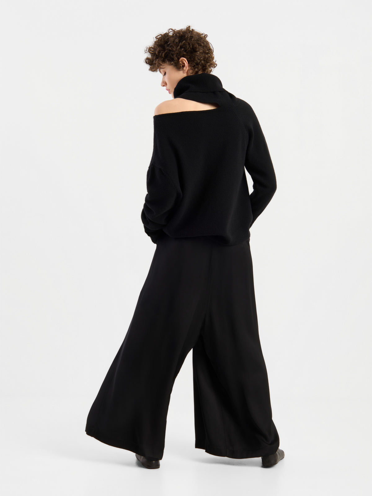 Eine Person mit kurzen Locken, die den Pullover „Scaya Schwarz“ trägt – einen locker sitzenden schwarzen Kaschmirpullover im schulterfreien Design –, kombiniert mit einer schwarzen Hose mit weitem Bein, steht mit dem Rücken zu einem schlichten weißen Hintergrund.