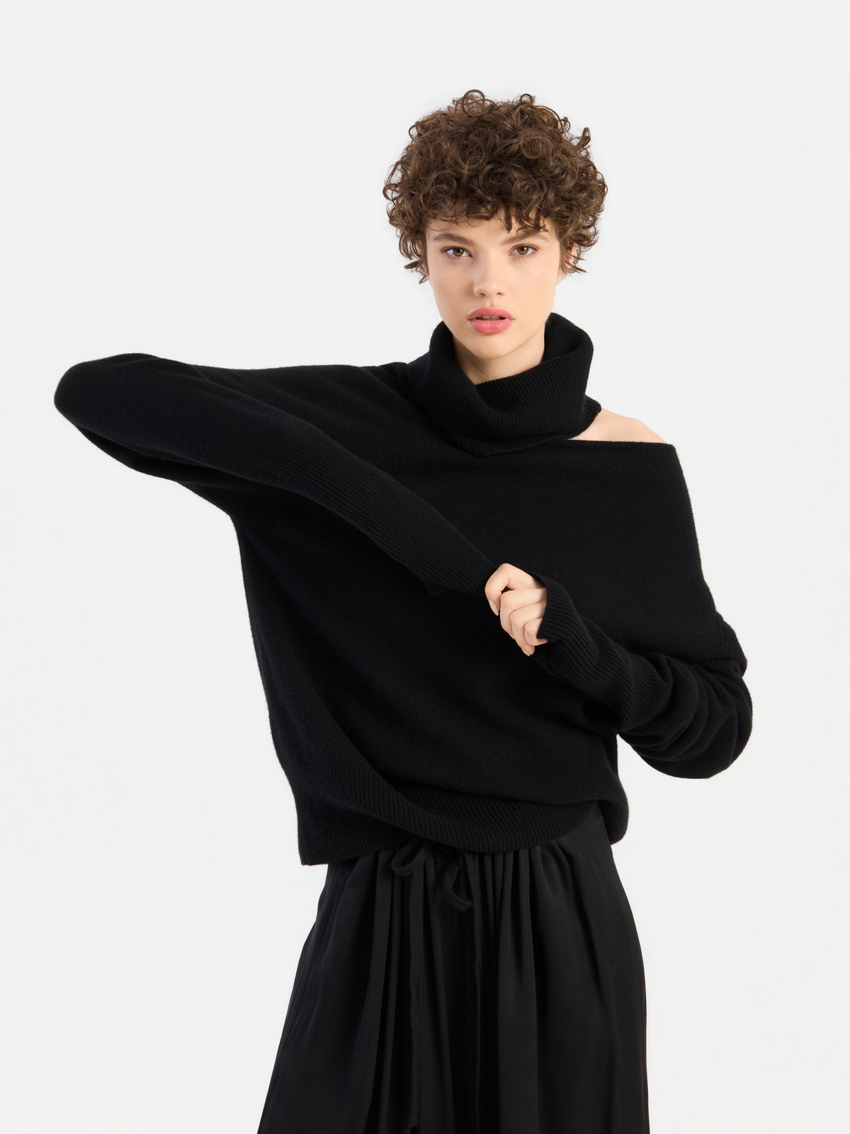Eine Frau mit kurzen Locken trägt den Pullover „Scaya Schwarz“, einen eleganten schwarzen Rollkragenpullover, und zieht ihren Ärmel zurecht, wobei sie einen Arm quer über den Körper hebt – vor einem schlichten weißen Hintergrund.
