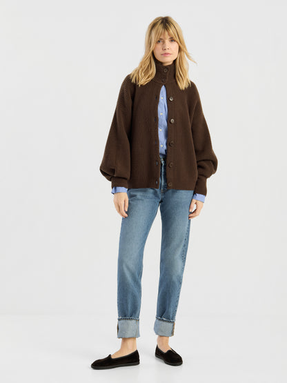 Vor einem schlichten Hintergrund steht eine Frau, die die Strickjacke „Bomber Braun“ über einem blauen Hemd, einer Jeans mit umgeschlagenen Hosenbeinen und schwarzen Loafern trägt. Sie hat blondes Haar und einen ausdruckslosen Gesichtsausdruck.