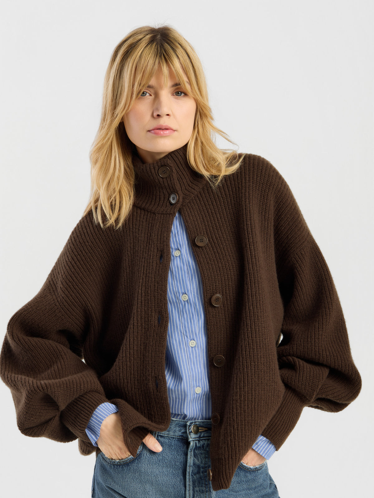 Eine Person mit blonden Haaren trägt die „Strickjacke Bomber Braun“, eine grob gestrickte Strickjacke aus weicher Merinowolle, die über einem blau gestreiften Hemd und einer blauen Jeans liegt, und steht mit den Händen in den Taschen vor einem schlichten, hellen Hintergrund.