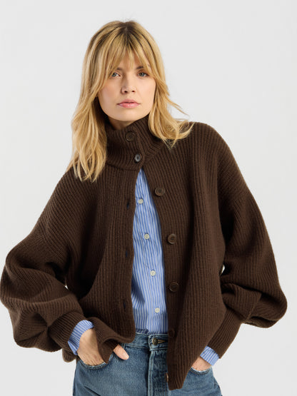 Eine Person mit blonden Haaren trägt die „Strickjacke Bomber Braun“, eine grob gestrickte Strickjacke aus weicher Merinowolle, die über einem blau gestreiften Hemd und einer blauen Jeans liegt, und steht mit den Händen in den Taschen vor einem schlichten, hellen Hintergrund.