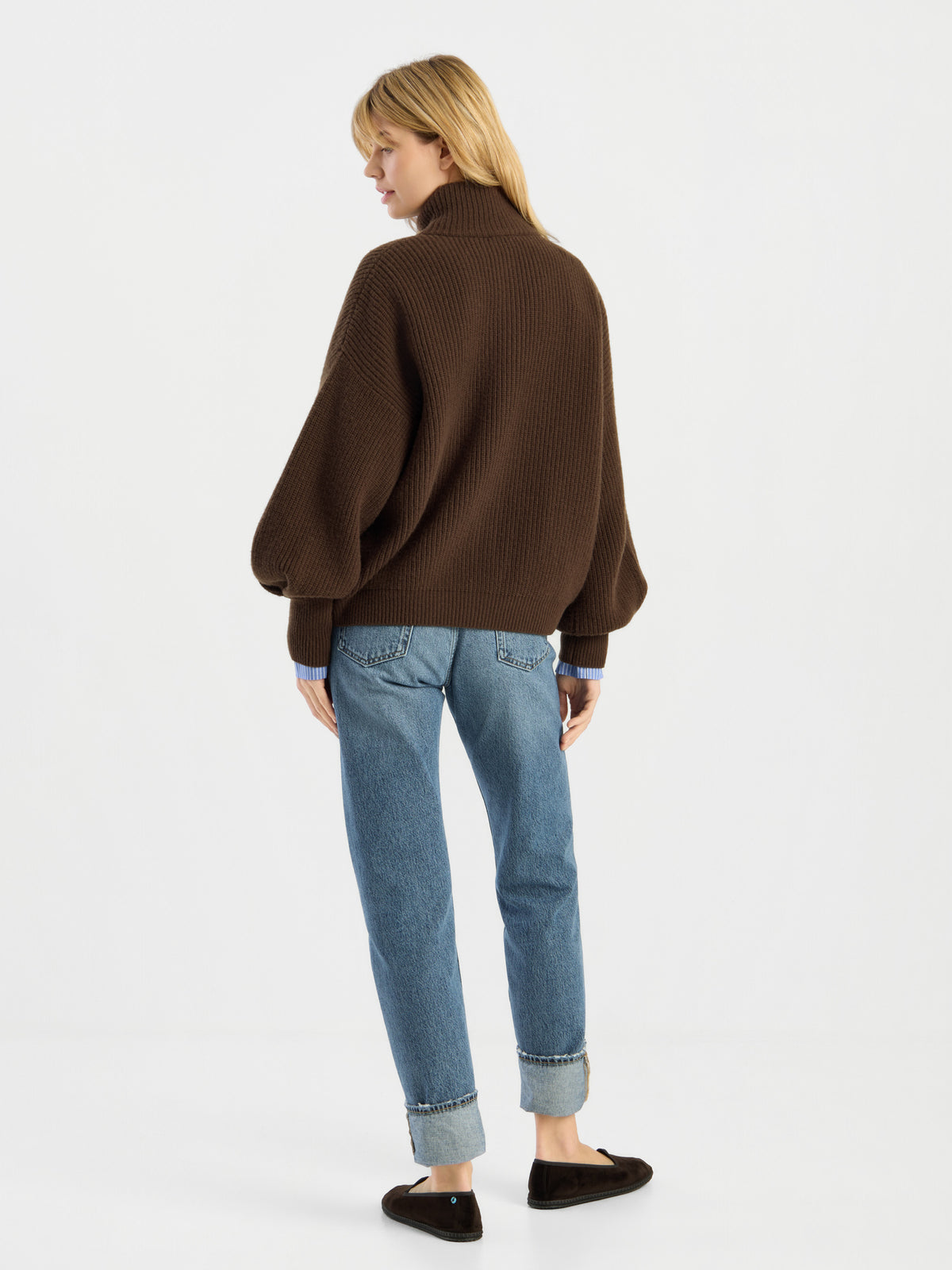 Eine Person steht vor einem weißen Hintergrund und trägt die Strickjacke „Bomber Braun“, eine blaue Jeans mit Umschlag und schwarze flache Schuhe; sie steht mit dem Rücken zur Kamera und hat den Kopf leicht zur Seite gedreht.