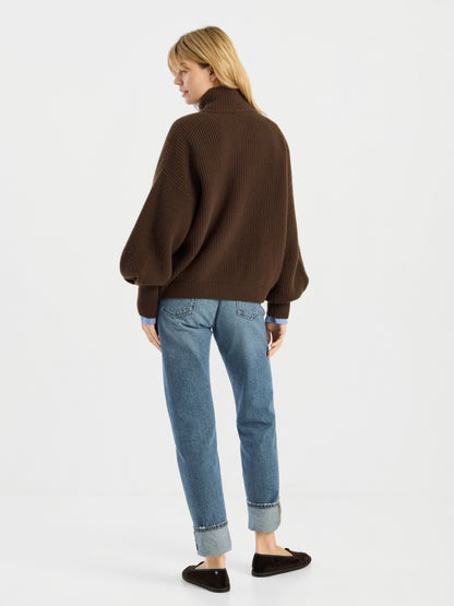 Eine Person steht vor einem weißen Hintergrund und trägt die Strickjacke „Bomber Braun“, eine blaue Jeans mit Umschlag und schwarze flache Schuhe; sie steht mit dem Rücken zur Kamera und hat den Kopf leicht zur Seite gedreht.