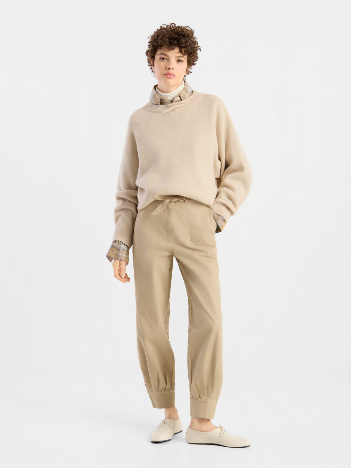 Eine Person steht vor einem schlichten weißen Hintergrund und trägt den beigen Pullover „Giulia Puder“ über einem karierten Hemd, dazu eine hellbraune Hose mit Umschlag und cremefarbene Schuhe. Sie hat kurzes, lockiges Haar und nimmt eine entspannte, selbstbewusste Haltung ein.