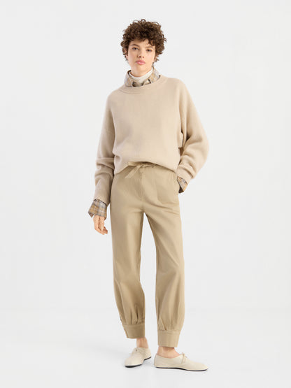 Eine Person steht vor einem schlichten weißen Hintergrund und trägt den beigen Pullover „Giulia Puder“ über einem karierten Hemd, dazu eine hellbraune Hose mit Umschlag und cremefarbene Schuhe. Sie hat kurzes, lockiges Haar und nimmt eine entspannte, selbstbewusste Haltung ein.