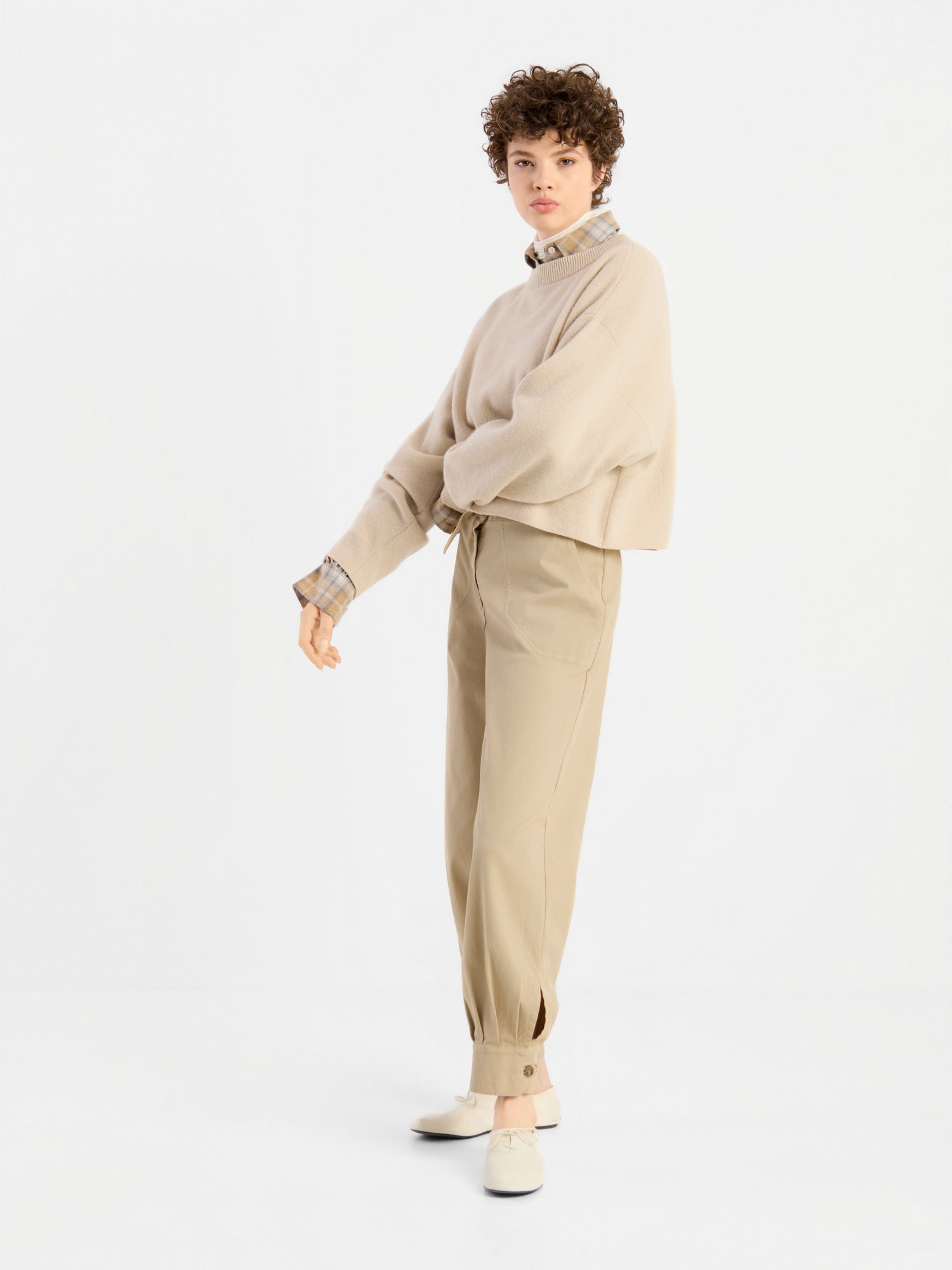 Eine Person mit kurzen Locken trägt einen beigen Pullover „Giulia Puder“ über einem karierten Hemd, eine khakifarbene Hose mit Knöpfen am Knöchel und beige Schuhe und steht vor einem schlichten weißen Hintergrund.