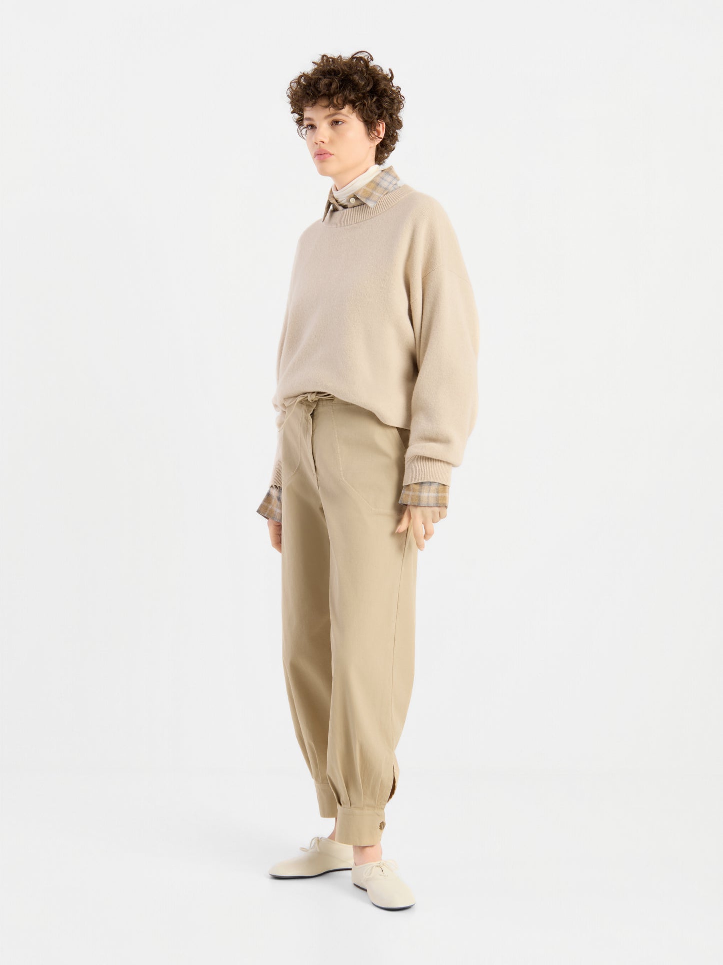 Eine Person mit kurzen Locken trägt den beigen Pullover „Giulia Puder“ über einem karierten Hemd, dazu eine lockere beige Hose und helle Schuhe, und steht vor einem schlichten weißen Hintergrund.