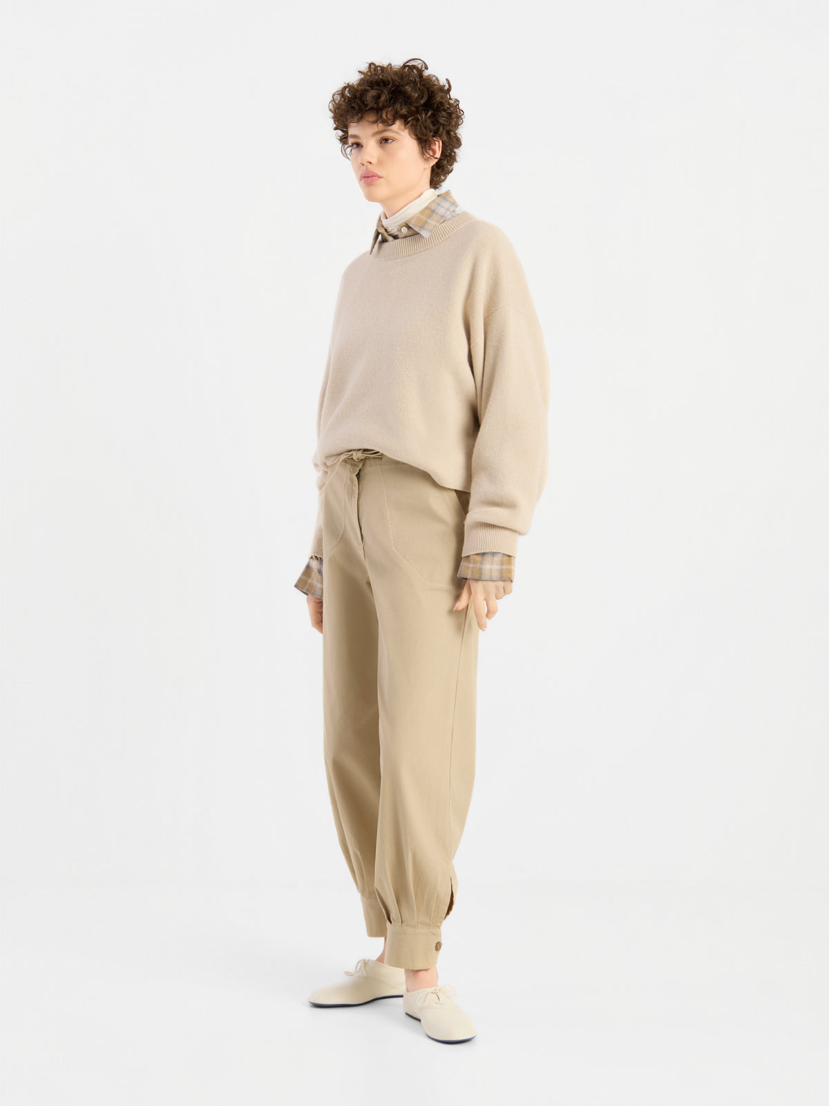 Eine Person mit kurzen Locken trägt den beigen Pullover „Giulia Puder“ über einem karierten Hemd, dazu eine lockere beige Hose und helle Schuhe, und steht vor einem schlichten weißen Hintergrund.