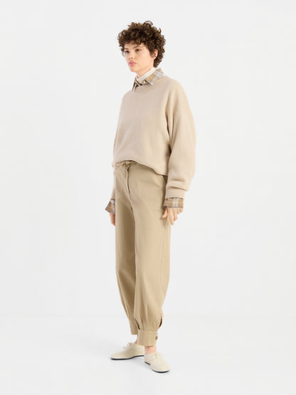 Eine Person mit kurzen Locken trägt den beigen Pullover „Giulia Puder“ über einem karierten Hemd, dazu eine lockere beige Hose und helle Schuhe, und steht vor einem schlichten weißen Hintergrund.