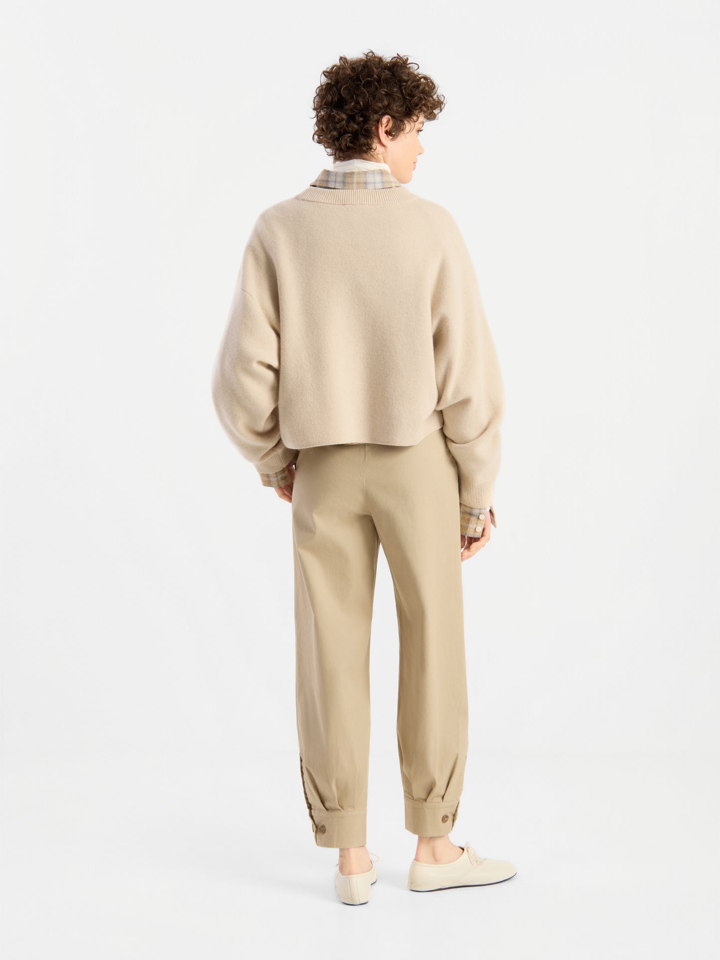 Eine Person mit kurzen, lockigen Haaren, die den Pullover „Giulia Puder“ aus Merinowolle über einem karierten Hemd trägt, dazu eine hellbraune Hose mit geknöpften Bündchen und beige Schuhe, steht mit dem Rücken zur Kamera in einem weißen Studio.