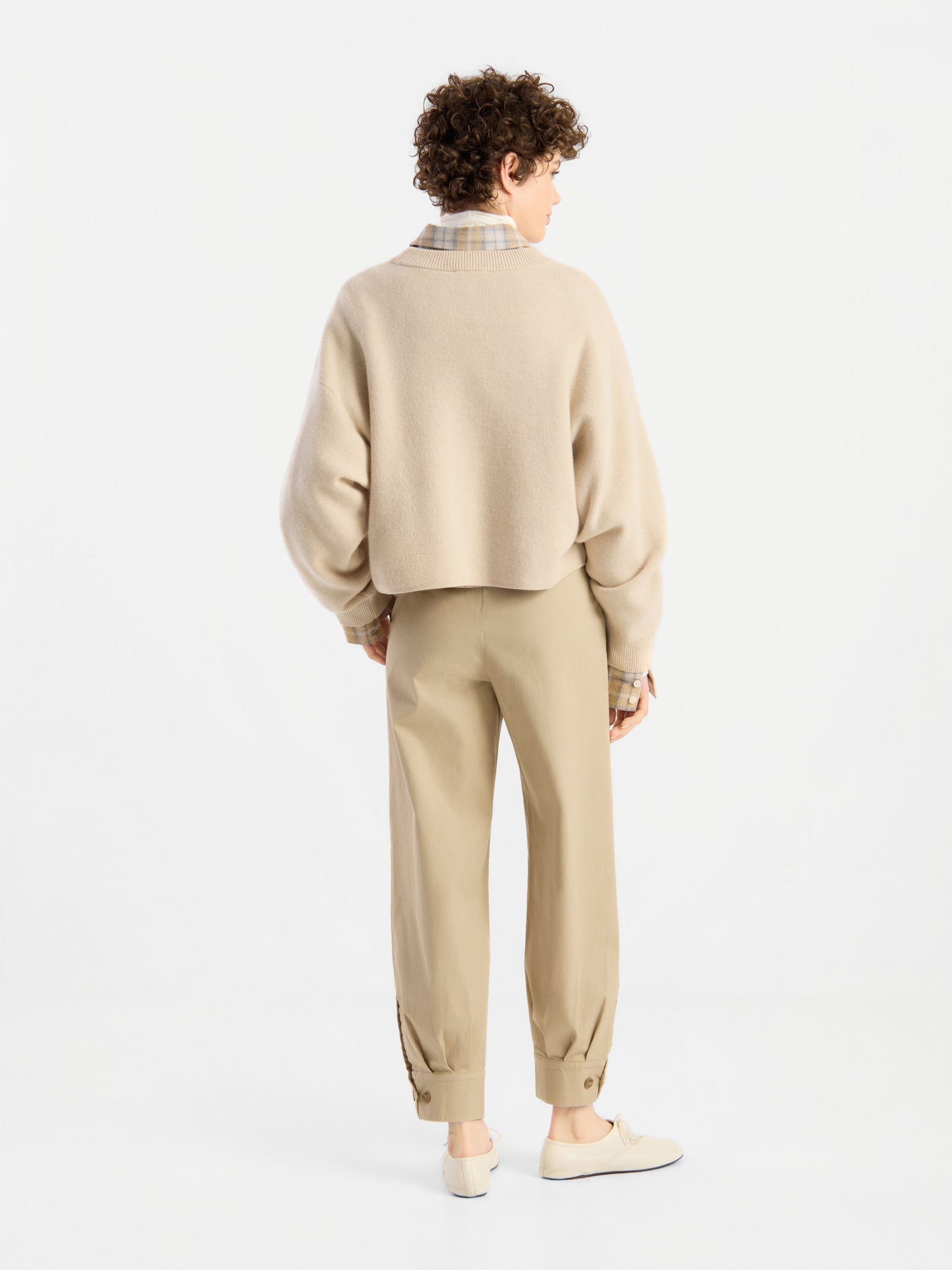 Eine Person mit kurzen, lockigen Haaren, die den Pullover „Giulia Puder“ aus Merinowolle über einem karierten Hemd trägt, dazu eine hellbraune Hose mit geknöpften Bündchen und beige Schuhe, steht mit dem Rücken zur Kamera in einem weißen Studio.